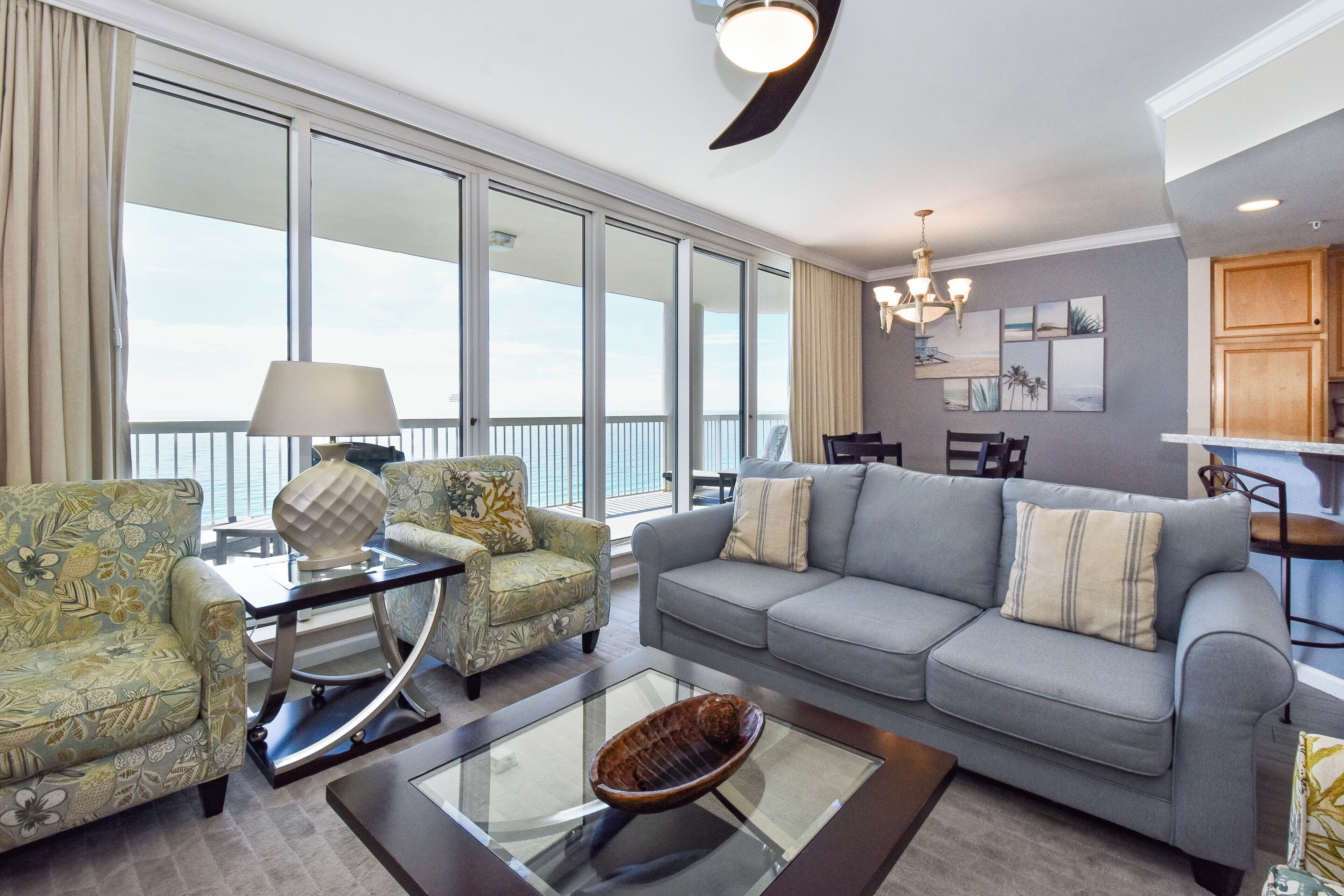 A spacious living room with a grey sofa, patterned armchairs, a glass coffee table, and a view through large windows.