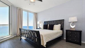A bedroom with a large bed, a nightstand, a lamp, and a view of the ocean through a sliding door. - A three-bedroom beachfront, modern Gulf Coast getaway! 🌴 (Destin)