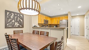 A dining area with a wooden table and chairs, a kitchen with yellow cabinets and stainless steel appliances, and a ceiling with recessed lighting. - Maui Resort Rentals: Westin Ka'anapali Ocean Resort 2BR Direct Oceanfront Villa (Lahaina)