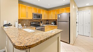 A kitchen with granite countertops, stainless steel appliances, wooden cabinets, and a white door. - Maui Resort Rentals: Westin Ka'anapali Ocean Resort 2BR Direct Oceanfront Villa (Lahaina)