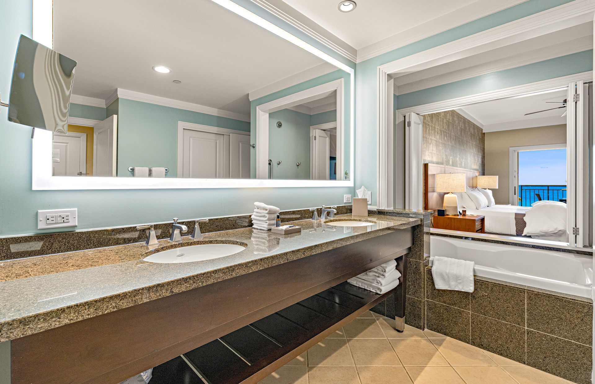A hotel bathroom with a double sink, a large mirror, and a view of a balcony.