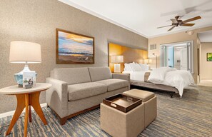 A hotel room with a sofa, a bedside table with a lamp, a bed with white bedding, a ceiling fan, and a painting on the wall.