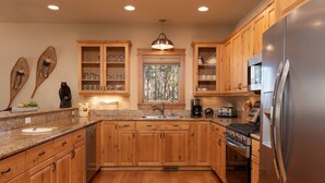 A kitchen with wooden cabinets, granite countertops, a stainless steel refrigerator, and a window with a view of trees.