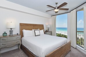 A bedroom with a large bed, a ceiling fan, a nightstand with a lamp, and a view of the beach.