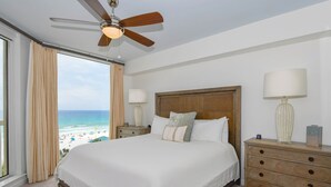 A bedroom with a large bed, a ceiling fan, a nightstand, a lamp, and a view of the beach. - Updated chic, modern beachfront condo w/private balcony! (Destin)
