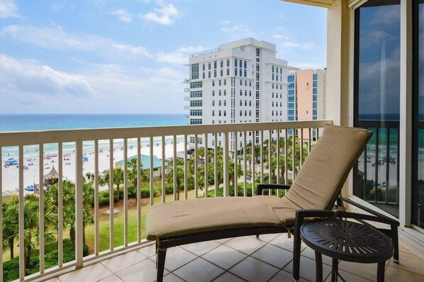 Ein Balkon mit Stuhl und Tisch mit Blick auf einen Strand, Palmen und ein Hotelgebäude im Hintergrund.