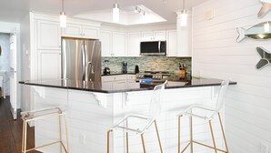 A modern kitchen with white cabinetry, stainless steel appliances, a black countertop, and three bar stools. - Updated chic, modern beachfront condo w/private balcony! (Destin)