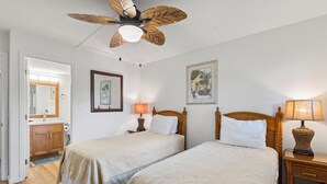 A bedroom with two beds, a ceiling fan, bedside lamps, a wooden headboard, a framed picture, and a bathroom visible through an open door. - Corner Beachfront Delight: Kihei Beach 110 Convenient Ground-Floor Haven! (Kihei)