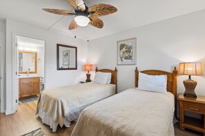 A bedroom with two beds, a ceiling fan, bedside lamps, a wooden headboard, a framed picture, and a bathroom visible through an open door. - Corner Beachfront Delight: Kihei Beach 110 Convenient Ground-Floor Haven! (Kihei)
