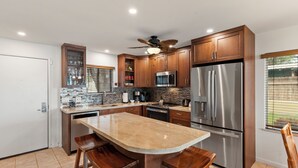 A modern kitchen with wooden cabinets, stainless steel appliances, a marble countertop, and a ceiling fan. - Corner Beachfront Delight: Kihei Beach 110 Convenient Ground-Floor Haven! (Kihei)