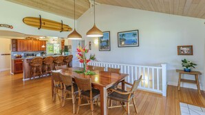 Una cocina amplia con gabinetes de madera, un comedor con mesa de madera y sillas de mimbre, y vista al océano a través de ventanas grandes.
