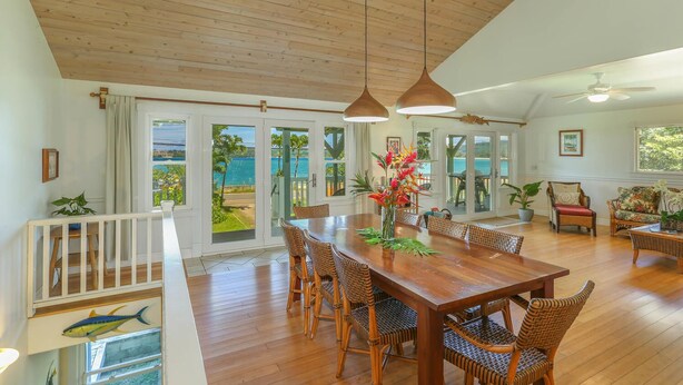 Un comedor espacioso con una mesa de madera y sillas de mimbre, ventanas amplias con vistas al mar y un techo de madera.