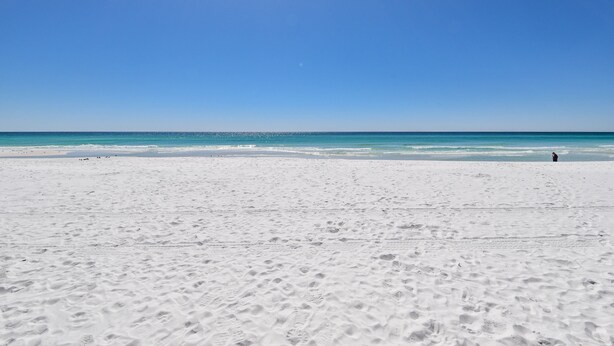 Une vaste plage de sable où l’on peut voir des empreintes de pas menant à l’eau, sous un ciel bleu dégagé.