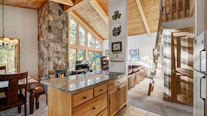 A spacious kitchen with a stone fireplace, wooden ceiling beams, and a large island with granite countertop. - Unwind in Alpine Bliss! River Views & Convenient Walkability to Town/Slopes (Breckenridge)