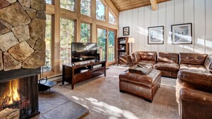 A spacious living room with a stone fireplace, leather sofas, a television, and large windows offering a view of the outdoors. - Unwind in Alpine Bliss! River Views & Convenient Walkability to Town/Slopes (Breckenridge)