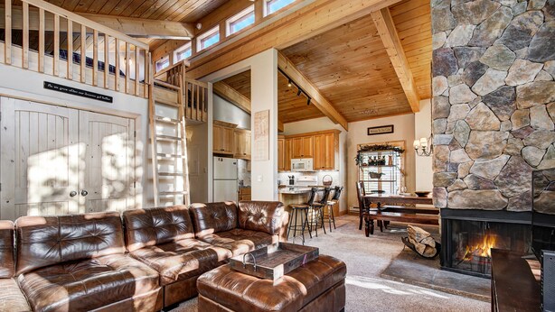 A spacious living room with a leather sectional sofa, a stone fireplace, and a kitchen in the background. - Unwind in Alpine Bliss! River Views & Convenient Walkability to Town/Slopes (Breckenridge)
