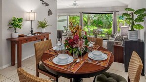 A dining area with a round table set for four, surrounded by wicker chairs. There is a wooden console table with potted plants and a lamp to the left. The room has a large window with a view of greenery outside. - Manualoha 1103 Garden View By Suite Paradise (Koloa)