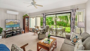 A living room with a television, sofa, coffee table, and a view of a tropical garden through large sliding glass doors. - Manualoha 1103 Garden View By Suite Paradise (Koloa)