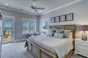 A bedroom with a bed, a nightstand, a ceiling fan, and a view of the beach through large windows.
