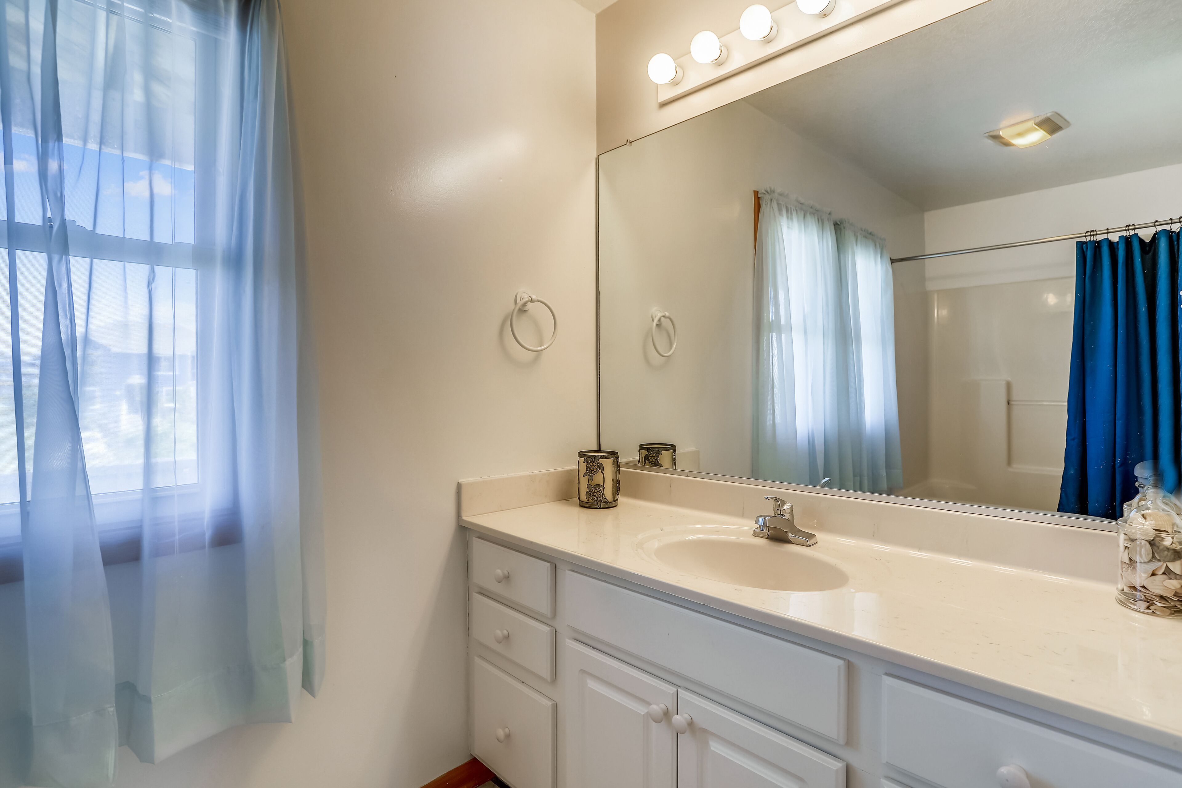 A bathroom with a large mirror, a sink, and a window with sheer curtains.