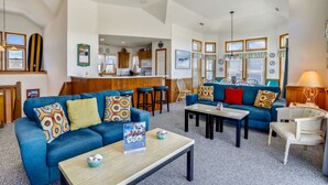Een ruime woonkamer met een blauwe zithoek, een salontafel en een eettafel. In de achtergrond is een keuken zichtbaar met houten kastjes en een bar.