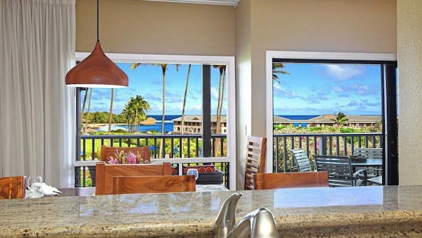 A kitchen with a view of a beach and palm trees, a dining area with wooden furniture, and a large window with a view of the ocean. - Poipu Sands 123 Beautiful Ocean View AC Suite Paradise (Koloa)