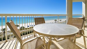 Un balcon agrémenté de fauteuils en osier et d’une table ronde, donnant sur une plage où l’eau est d’un bleu limpide et le rivage est sablonneux.