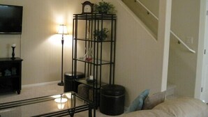 A living room with a glass coffee table, a black metal shelf with decorative items, a lamp, and a staircase.