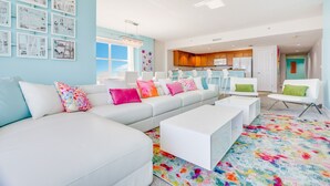 A modern living room with a white sectional sofa, colorful pillows, a vibrant rug, and a kitchen in the background.
