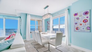 A bright room with a white dining table, chairs, and a chandelier. There are large windows with curtains, a white sofa with colorful pillows, and a painting on the wall.