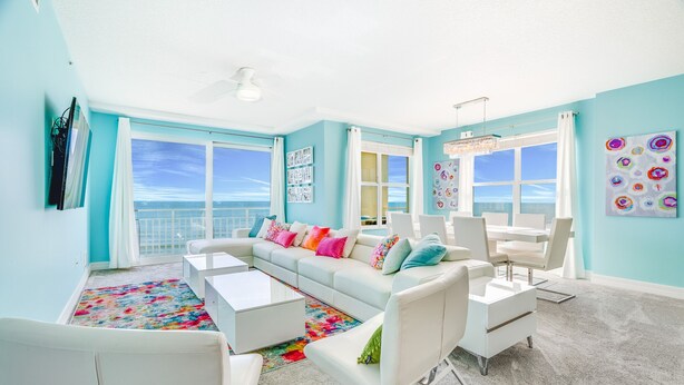 A bright living room with a white sofa, colorful pillows, a white coffee table, and a view of the ocean through large windows.