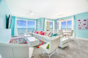 Un salon lumineux avec un canapé blanc, des coussins colorés, une table basse blanche et une vue sur l’océan grâce à de grandes fenêtres.