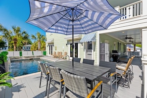A poolside area with a table and chairs, a large umbrella, and a view of a house with a balcony.