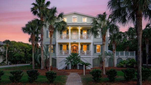 A two-story house with a front porch, a staircase leading to the entrance, and a well-maintained garden with palm trees and shrubs.