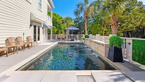 A swimming pool with lounge chairs and a pergola, surrounded by palm trees and a white house.