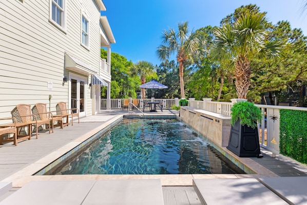A swimming pool with lounge chairs and a pergola, surrounded by palm trees and a white house.