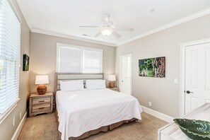 A bedroom with a bed, two bedside tables with lamps, a ceiling fan, a window with blinds, and a door.