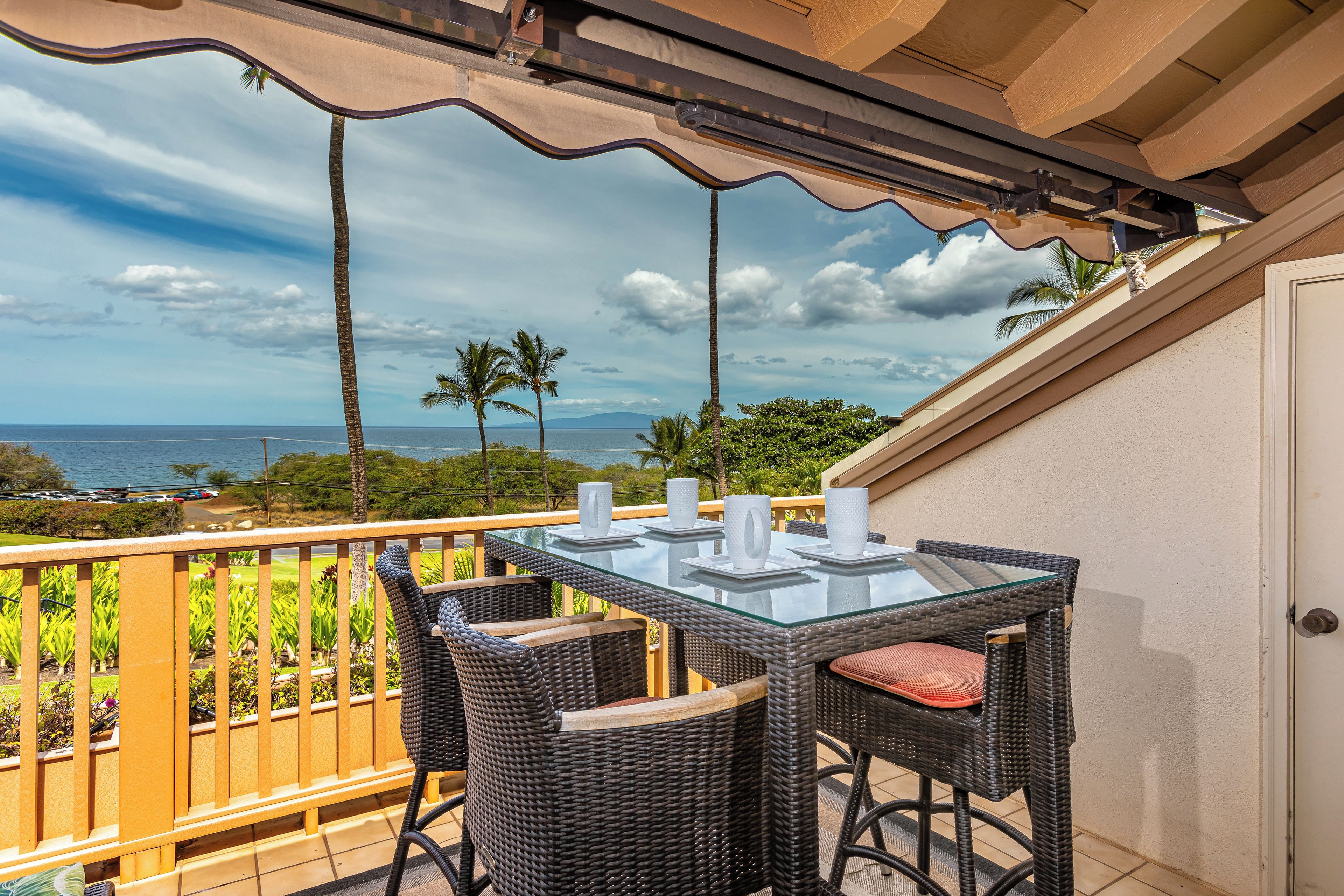 A balcony with a glass table and wicker chairs, overlooking a scenic ocean view with palm trees and a clear sky.
