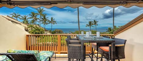 A covered patio with a table and chairs, overlooking a view of palm trees and the ocean.