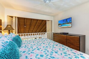A bedroom with a wooden ceiling, a bed with a floral bedspread, a nightstand with a lamp, a dresser, and a TV displaying a beach scene.