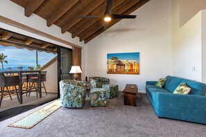 A spacious living area with a blue sofa, patterned ottomans, a wooden coffee table, and a dining set. A ceiling fan and a painting of a tropical scene are also visible.