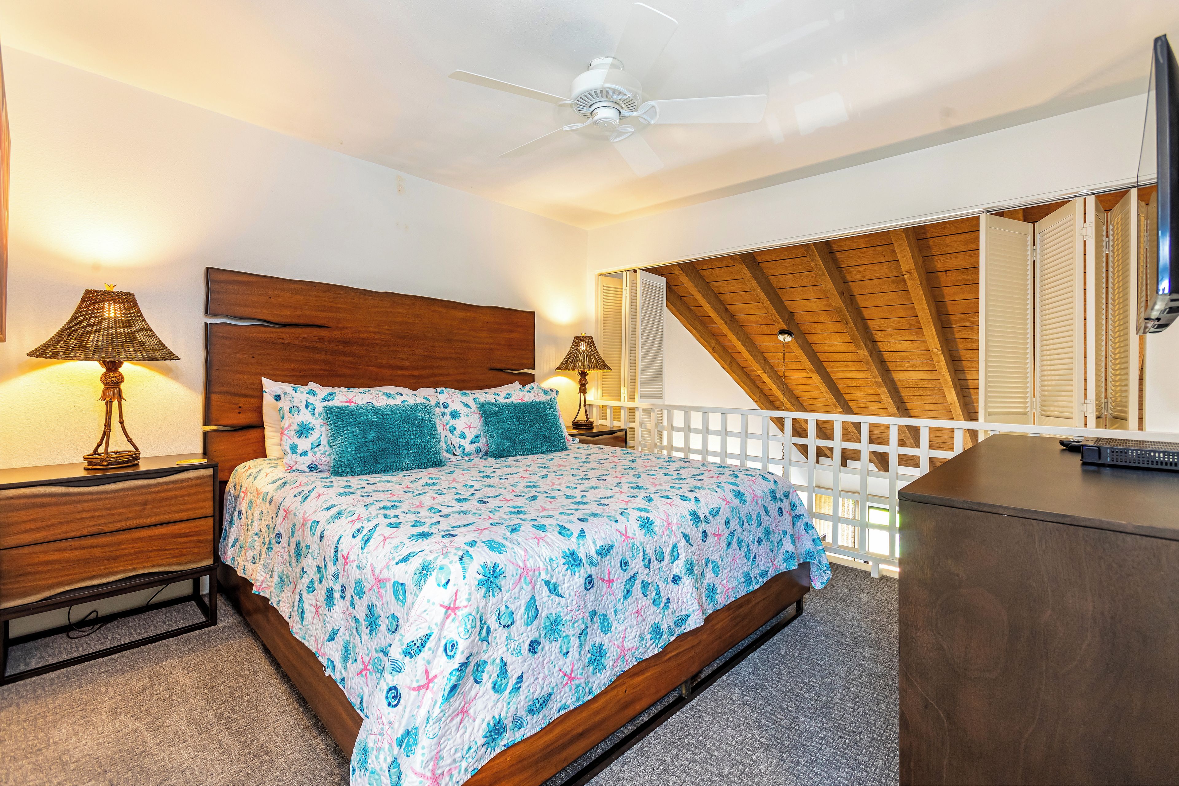 Een slaapkamer met een houten hoofdbord, een bed met een bloemenbedekking, twee bedlampen, een plafondventilator en een televisie op een houten dressoir.