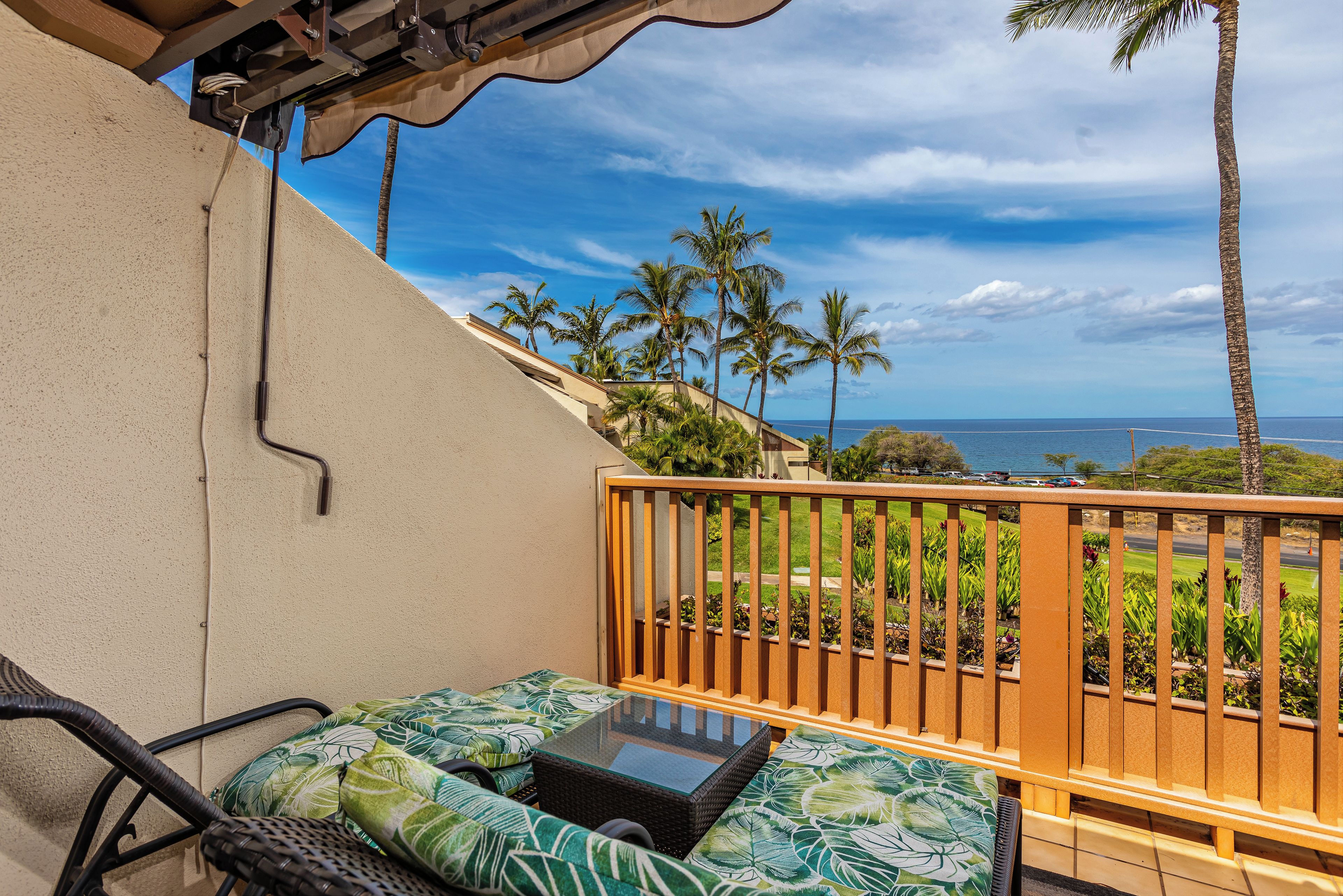 A balcony with a glass table, wicker chairs, and a view of the ocean and palm trees.