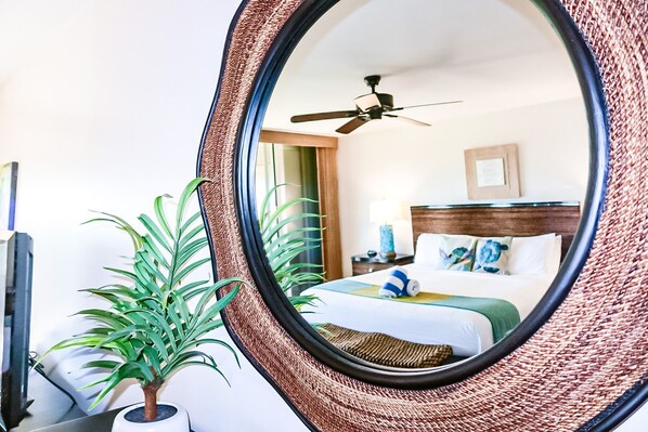 Une chambre d’hôtel avec un grand miroir, un ventilateur de plafond, un lit recouvert d’une courtepointe verte et une plante en pot.