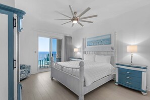 A bedroom with a white bed, a ceiling fan, a nightstand with a lamp, a blue dresser, and a view of the ocean through a sliding door.