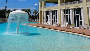 A swimming pool with a water fountain, surrounded by lounge chairs and a building with large windows and columns.