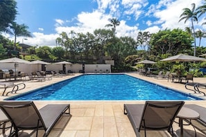 Une piscine entourée de chaises longues et de parasols, bordée de palmiers et surplombée d’un ciel dégagé.