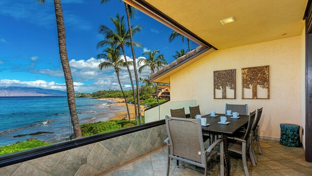 A balcony with a view of the beach and ocean, a dining table with chairs, and two wall-mounted artworks. - Beachfront Living - Makena Surf E-202 - Mesmerizing Views & Tropical Landscapes (Makena, Maui)