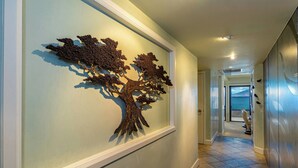 A hallway with a wall-mounted tree sculpture, a tiled floor, and a door leading to a room with a view. - Beachfront Living - Makena Surf E-202 - Mesmerizing Views & Tropical Landscapes (Makena, Maui)
