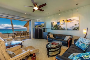 A spacious living room with a large window overlooking the ocean, a ceiling fan, and a flat-screen TV.
